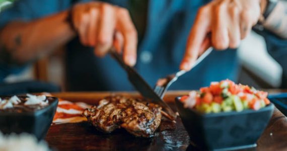 Hands holding fork and knife and eating delicious juicy steak on a wooden plate
